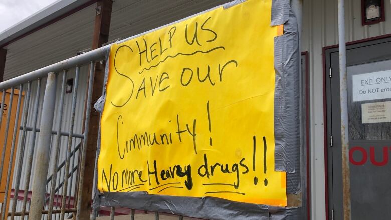 A handmade sign taped to the railing outside a building reads, 'Help us save our community! No more heavy drugs.' 