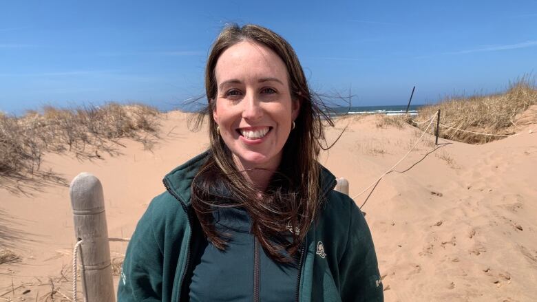 A brown haired woman in a green jacket stands in front of a beach