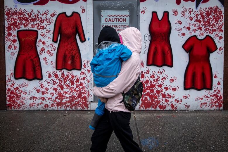 A passerby in a raincoat, carrying a small child, walks by mural paintings of red dresses.