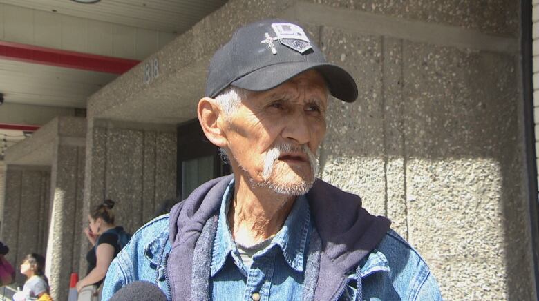 A man in a hat stands behind several news microphones speaking to reporters outside a downtown hotel in broad daylight.