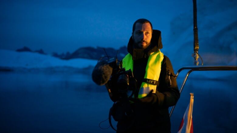 A man wearing a black jacket and a yellow safety vest holds a video camera while standing outside at dusk.