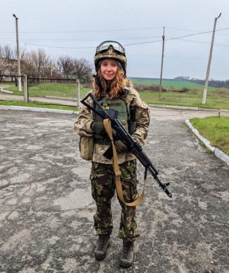 A woman with curly hair wearing military fatigues, a helmet and holding a gun, stands on an empty street.