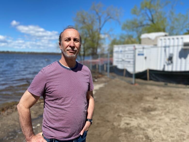 A man in a striped shirt stands on a sandy beach. The Ottawa River is in the background