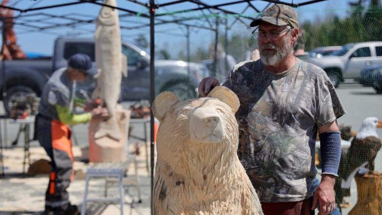 An artist stands in front of a wooden bear he carved with a chainsaw. 