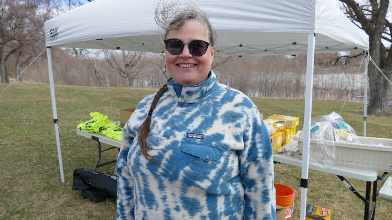 A woman in a blue and white sweater smiles at the camera as she stands in a park near a river, with a table covered with T-shirts and cleaning supplies behind her.