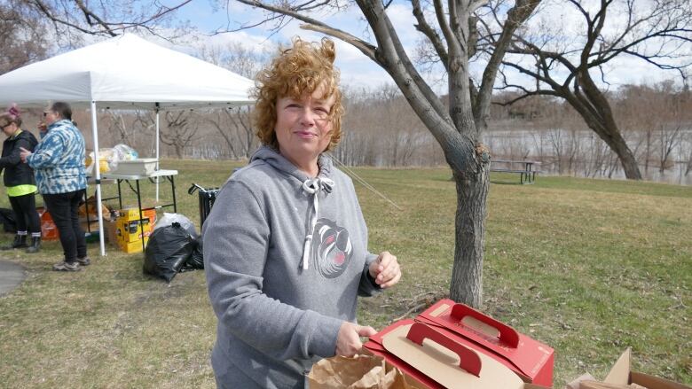 A woman in a grey hoodie smiles at the camera as she stands in a park near a river, with a table covered with T-shirts and cleaning supplies behind her