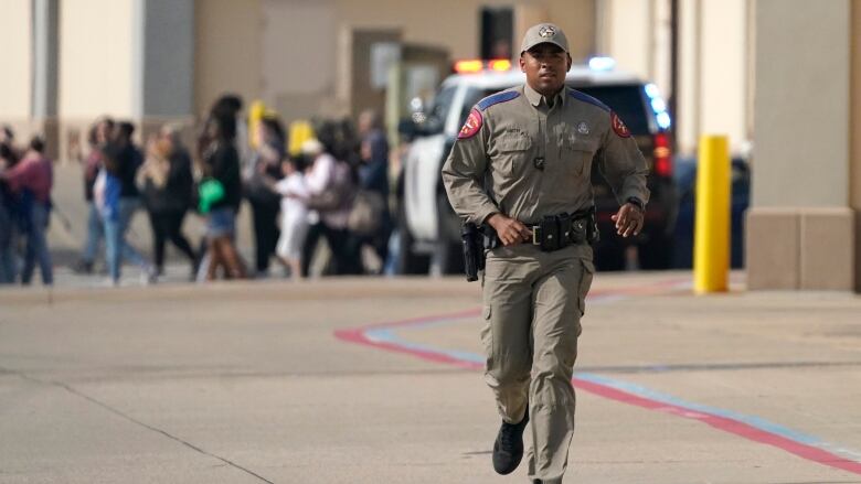 A law enforcement officer runs toward the camera as people are seen walking in a line in the background.