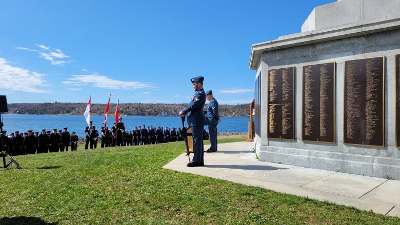 A man in uniform stands with a gun for display by a monument with more uniformed members in the background and the ocean.