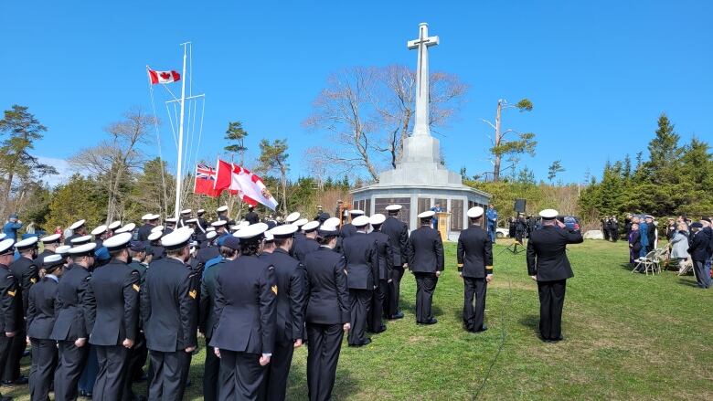 Men and women in uniform salute to a memorial with a cross on the top of it.