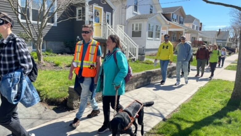 A woman with a guide dog walks along a city pavement followed by a group of other people