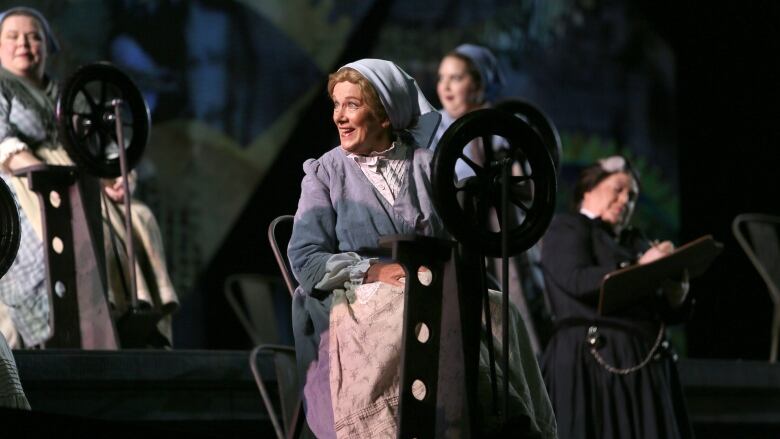 A woman in a costume from Richard Wagner's The Flying Dutchman opera sings on stage with other performers around her.