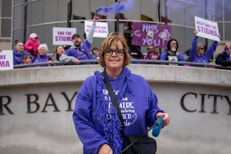 A woman wearing purple stands outside, smiling, with a crowd of people standing behind her.