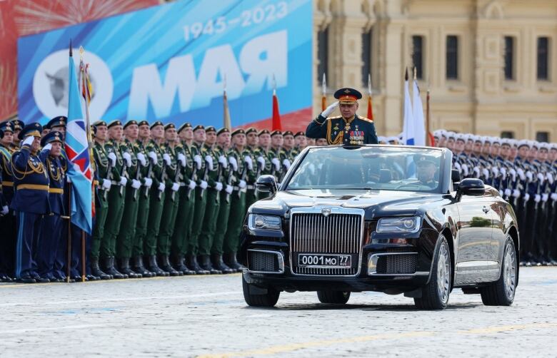 A man in a convertible vehicle stands and salutes during a parade, with soldiers seen standing along the side of the road.