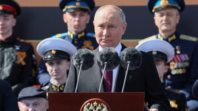 A man is shown standing at a podium with microphones in an outdoor photo.