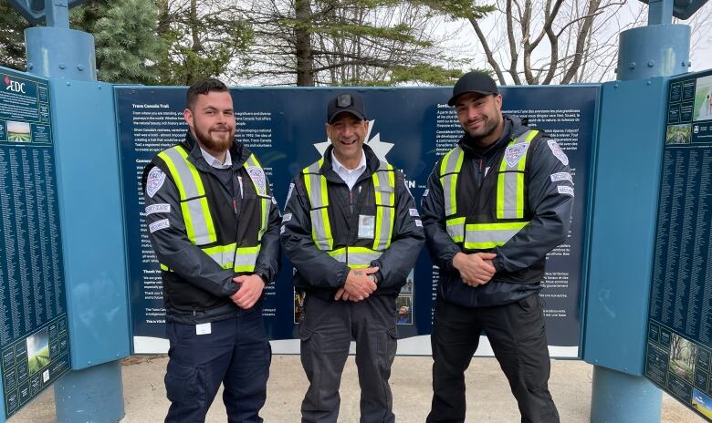 Three uniformed men pose together in front of a trail sign. 