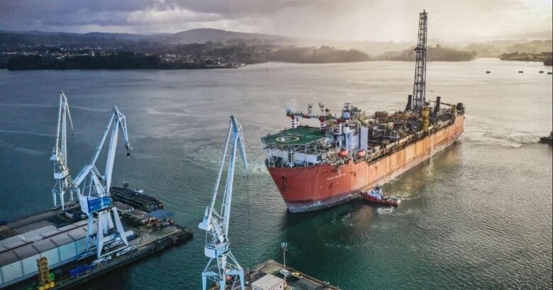  A 300-metre rusty ship being assisted by tugboats leading it into a port with cranes. 