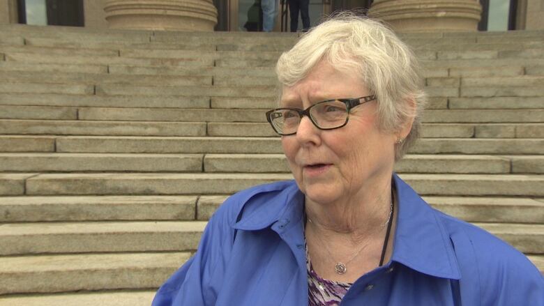 A woman with short white hair and glasses wearing a blue top stands against steps and looks off to the side.