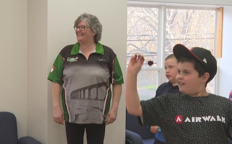 10 year old William Wright smiles in a black shirt and cap as he gets ready to throw his dart while coach Tracy Power smiles in the background. 