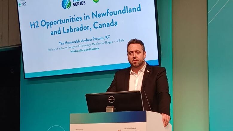 A man in a suit speaking at a podium with a screen behind him that says 