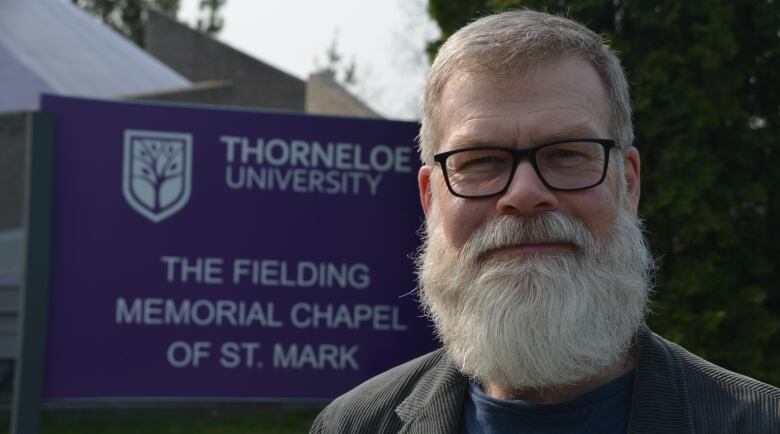 A man with a long white beard stands in front of a Thorneloe University sign 