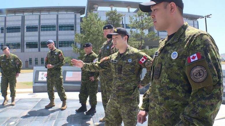 Several Canadian soldiers stand in an open space with trees and a building in the background.