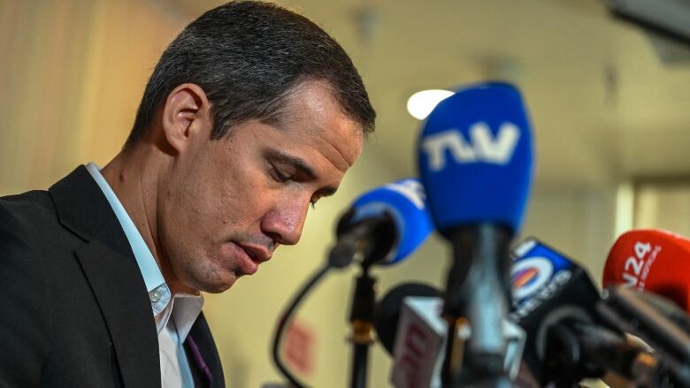 Juan Guaido, a man with close-cropped hair wearing an open-collared shirt and suit jacket, sits in front a set of microphones with his eyes closed. 