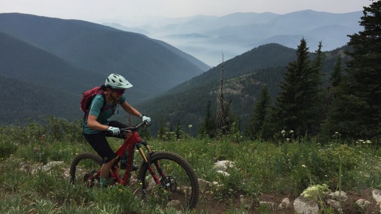 A woman wearing a helmet is seen riding a mountain bike on a trail. Mountains can be seen in the background.