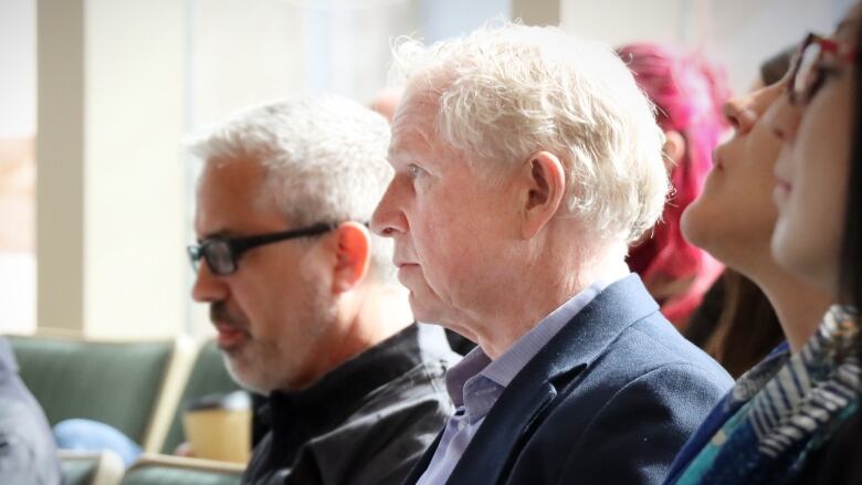 A man with white hair and a blue suit in the middle of some other people listens as someone else is speaking.