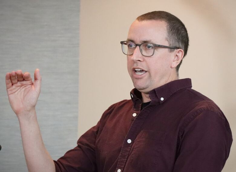 A man with a red shirt and glasses gestures with his hand as he speaks.