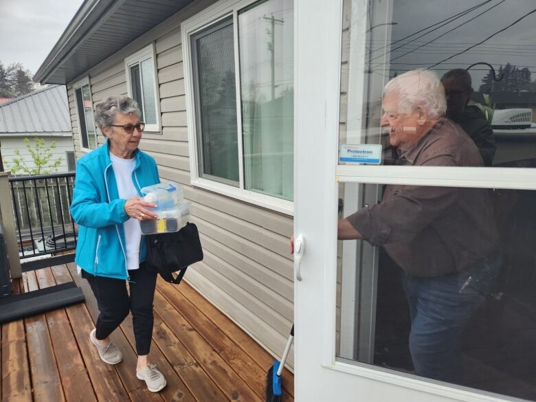 A grey-haired woman with a blue jacket and sunglasses carries something into her home where a white-haired man stands holding the door. 