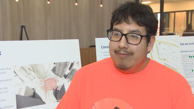 A man wearing an orange shirt and glasses is standing in front of boards displaying words and images.