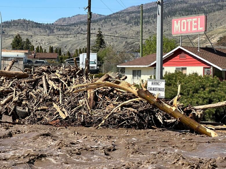 Wood debris lines a flowing river, with a motel and hills in the background.