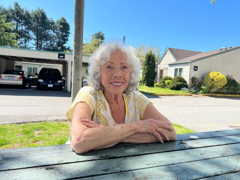 A woman with grey hair and a yellow shirt sits at a park table outside. Behind her is a road and some houses. 