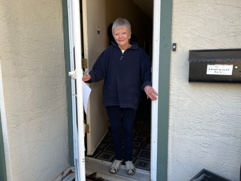 A woman with grey hair and a blue jacket stands in a doorway with the door open. 