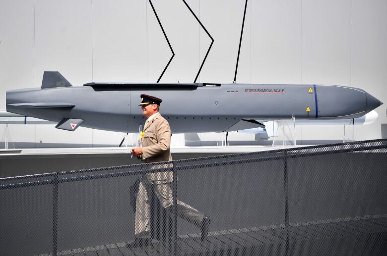 A man in a military-style cap walks down a ramp path in front of a missile display.