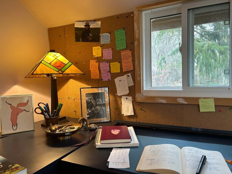 A black desk against a wall with a window and cork board with a lamp and notebooks on it and a writing journal