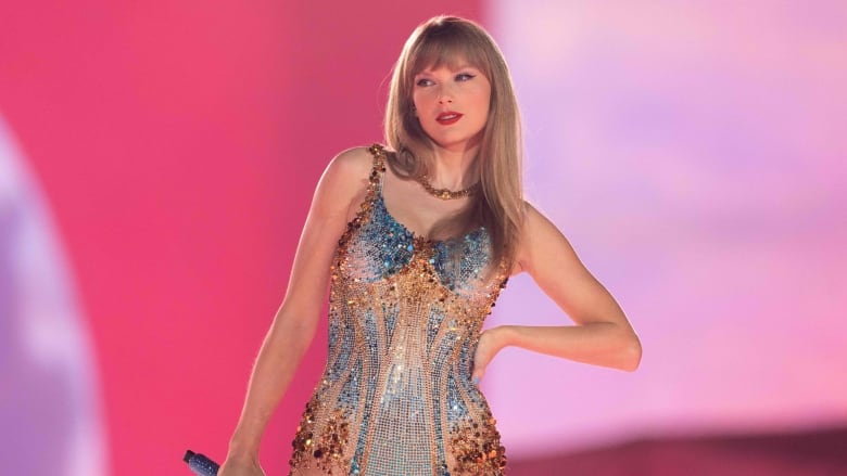 A blond woman in a sparkly gold and blue leotard puts her hand on her hip, the other hand holding a microphone.