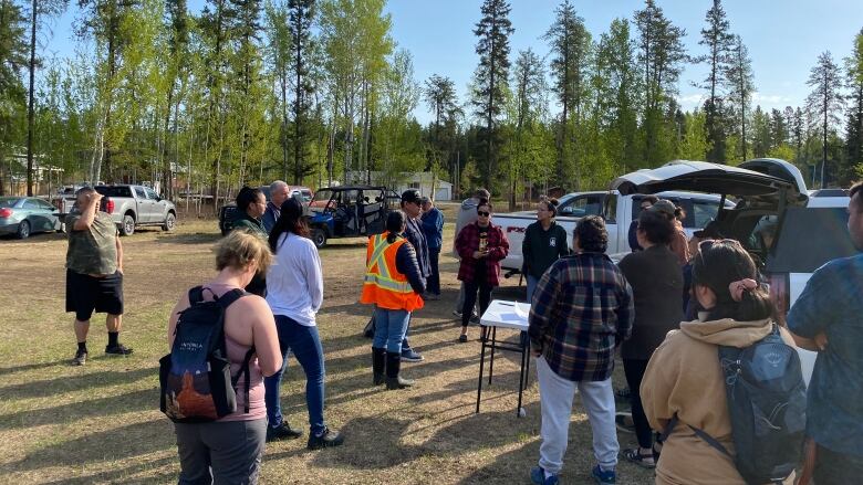 Seventeen people gather outside. There are parked cars and trees in the background.