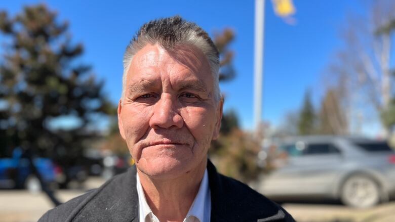 An Indigenous man with salt and pepper hair stands in the sun in front of an orange flag