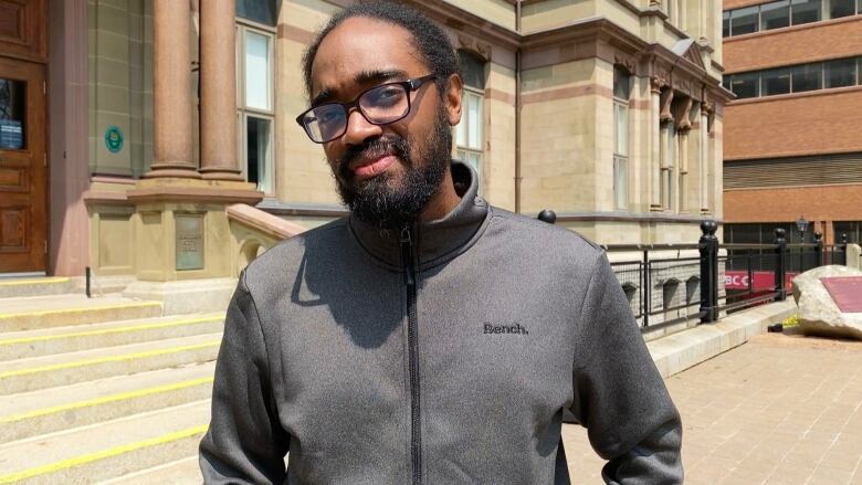 A Black man with a black beard and glasses stands outside Halifax city hall, wearing a grey sweater