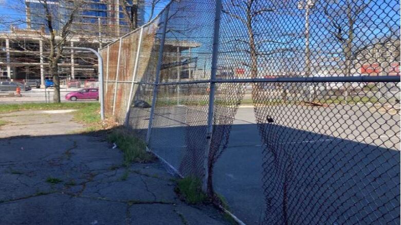 A chainlink fence is shown with a rip in it surrounding an overgrown cement area