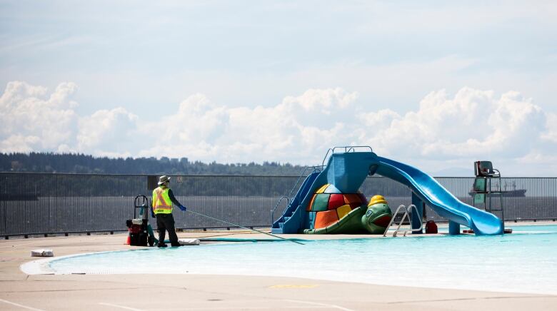 A worker in a yellow vest extends a wand into a pool near a waterslide.