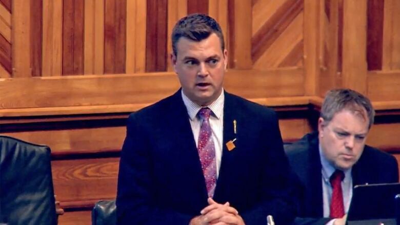 A man wearing a blazer, collared shirt and tie, standing up, speaking.