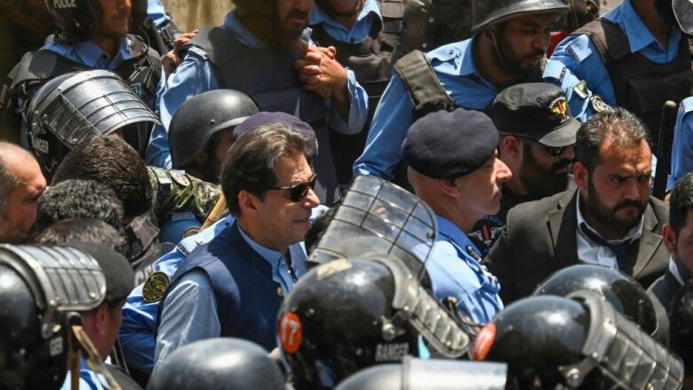 A man wearing sunglasses, a light blue shirt and dark blue vest is escorted into a court in Islamabad, Pakistan, by police officers, some wearing riot helmets.