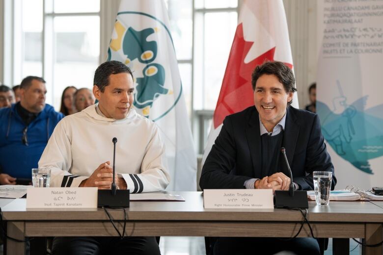 Two men sit at a table with flags and an audience in the background. 