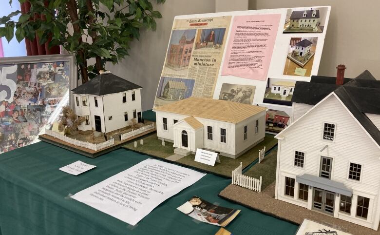 Three miniature buildings on a table covered in a green tablecloth. A poster board with newspaper clippings and photos leans on the wall behind the replicas.