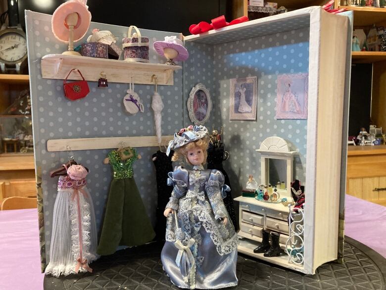 A doll wearing a blue gown in front of an open book box with a miniature vanity on the right and hanging dresses on the left.