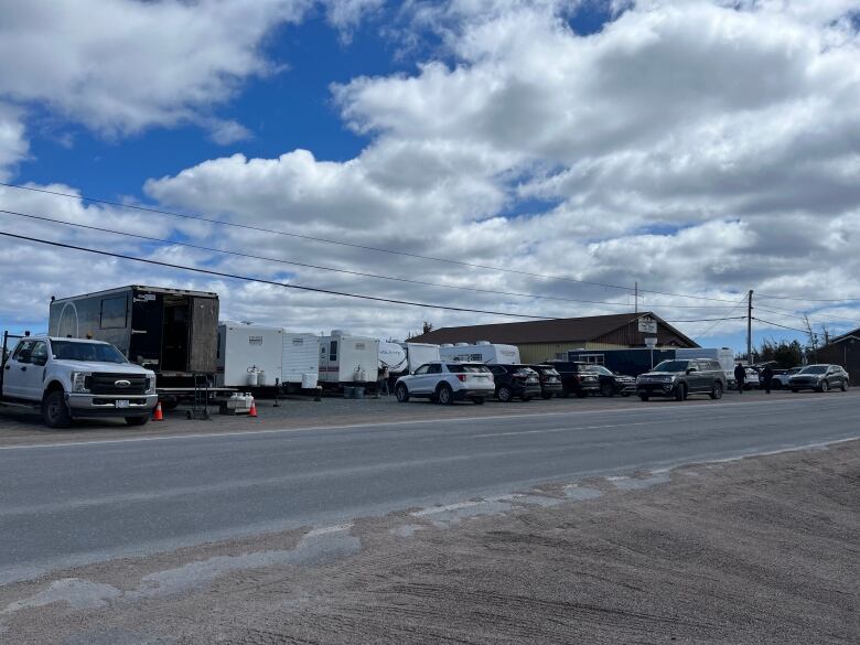 A large truck, several trailers, RVs, and other vehicles crowd a dirt parking lot, with a big blue sky above. 