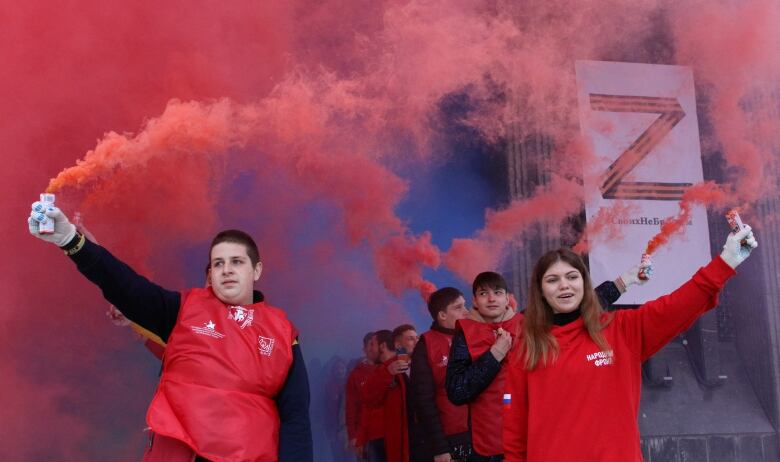 A group of young people wearing red hold smoke flares, while a board displays the symbol Z.