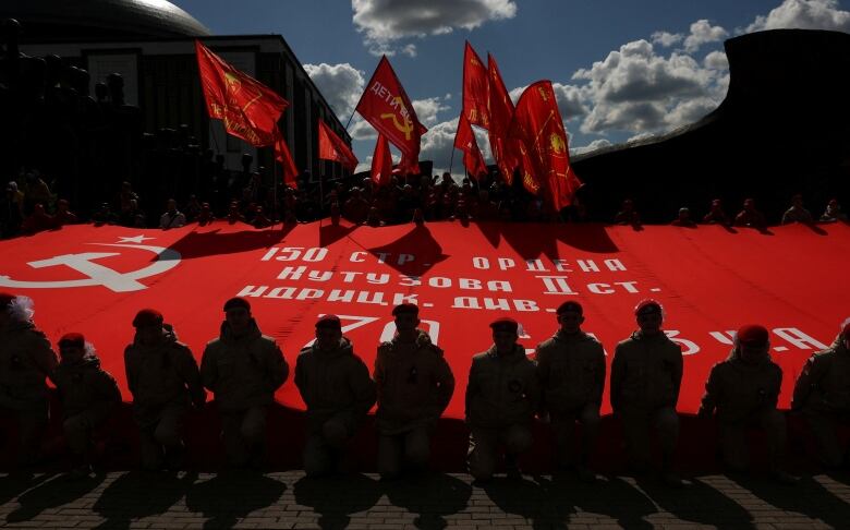 People wearing military uniforms, some holding flags, surround a large red banner.